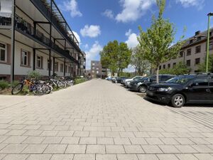 Eine gepflasterte Straße, links davon Gebäude, rechts Parkplätze